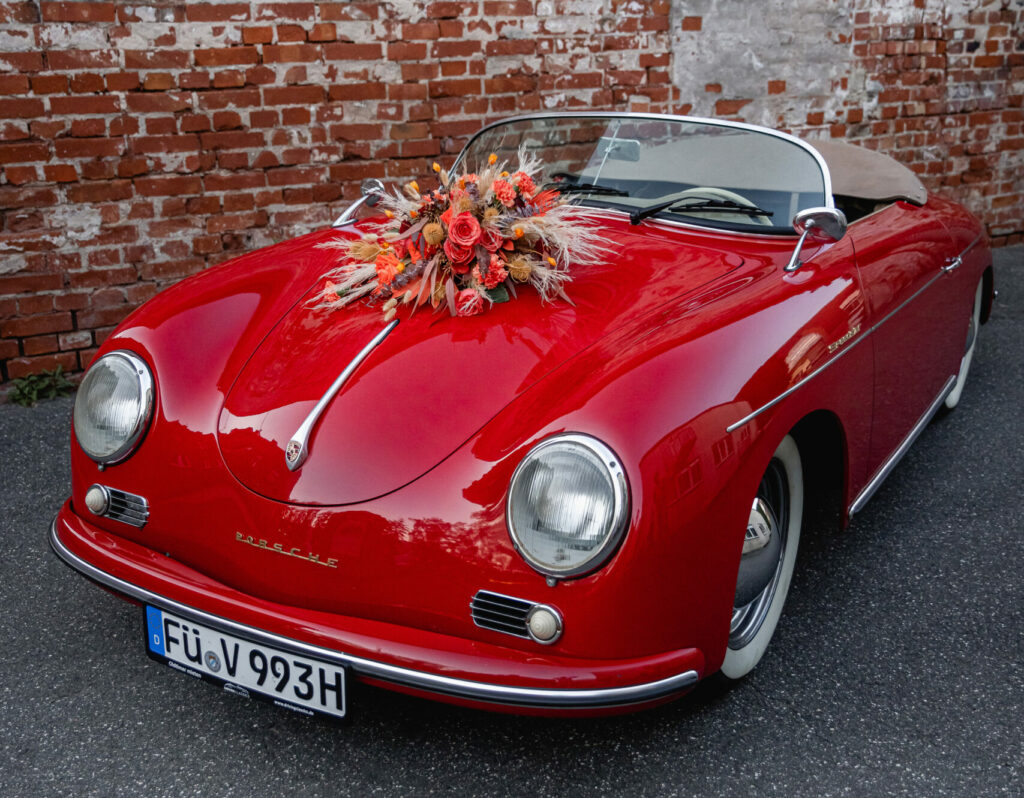 Porsche 356 Speedster in Rot mit Blumenschmuck auf der Haube neben Gemäuer