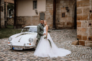 Hochzeitsauto Porsche 356 Coupé cremeweiß neben Brautpaar vor Gemäuer